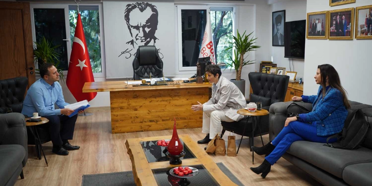Bornova Belediyesi'ndeki bir makam odasında, ahşap bir çalışma masasının etrafında üç kişi oturuyor. Masanın arkasında Mustafa Kemal Atatürk'ün silüeti ve Türk bayrağı yer alıyor. Mavi gömlekli bir erkek elinde dosyalarla masanın bir ucunda otururken, karşısında açık renkli kıyafetler giymiş bir kadın ve yanında mavi ceketli başka bir kadın daha oturuyor. Ortadaki ahşap sehpanın üzerinde kırmızı dekoratif objeler bulunuyor.