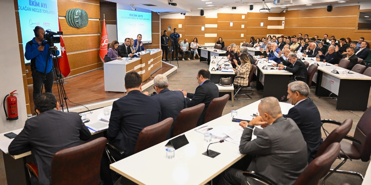 Bornova Belediyesi'nin Ekim ayı olağan meclis toplantısının yapıldığı geniş bir toplantı salonu. Salonda kürsüde oturan yetkililer ve sıralarda yerlerini almış meclis üyeleri bulunuyor. Bir kameraman toplantıyı kaydederken, katılımcıların bir kısmı ellerini kaldırarak oylamaya katılıyor. Salonun ön tarafında üzerinde 