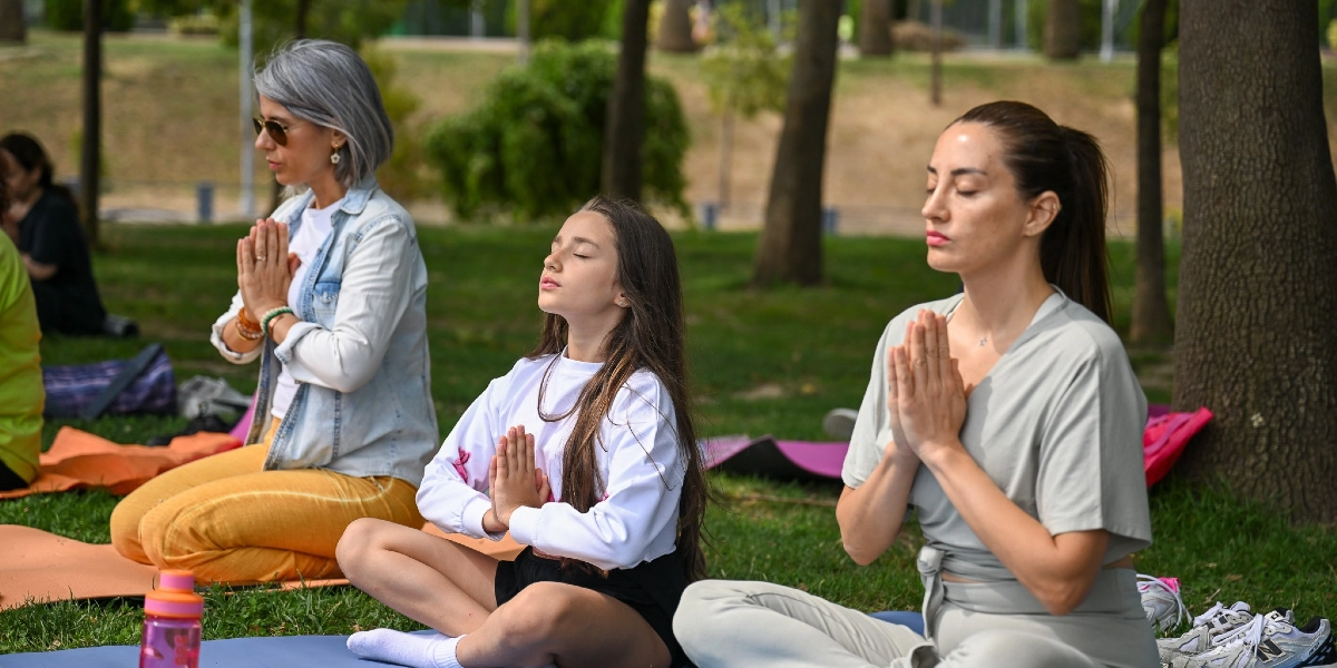 Ağaçlık bir park alanında, yoga matları üzerinde oturarak meditasyon yapan üç kadın ve bir kız çocuğu. Hepsi gözleri kapalı, ellerini göğüs hizasında birleştirmiş şekilde huzurla oturuyor. Ön planda gri kıyafetli bir kadın, yanında genç bir kız ve sol tarafta kot ceketli, sarı pantolonlu başka bir kadın yer alıyor. Arka planda ise yeşil çimenler ve ağaçlar görülüyor.