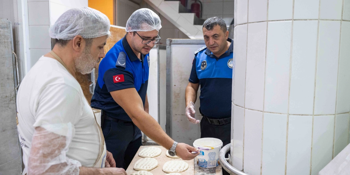 Beyaz önlük ve saç bonesi takan bir fırın çalışanı, tezgah üzerindeki hamurları hazırlarken, mavi üniformalı iki zabıta memuru denetim yapıyor. Zabıta memurlarından biri elindeki fırçayla hamurların üzerine sürülen malzemeyi kontrol ediyor.