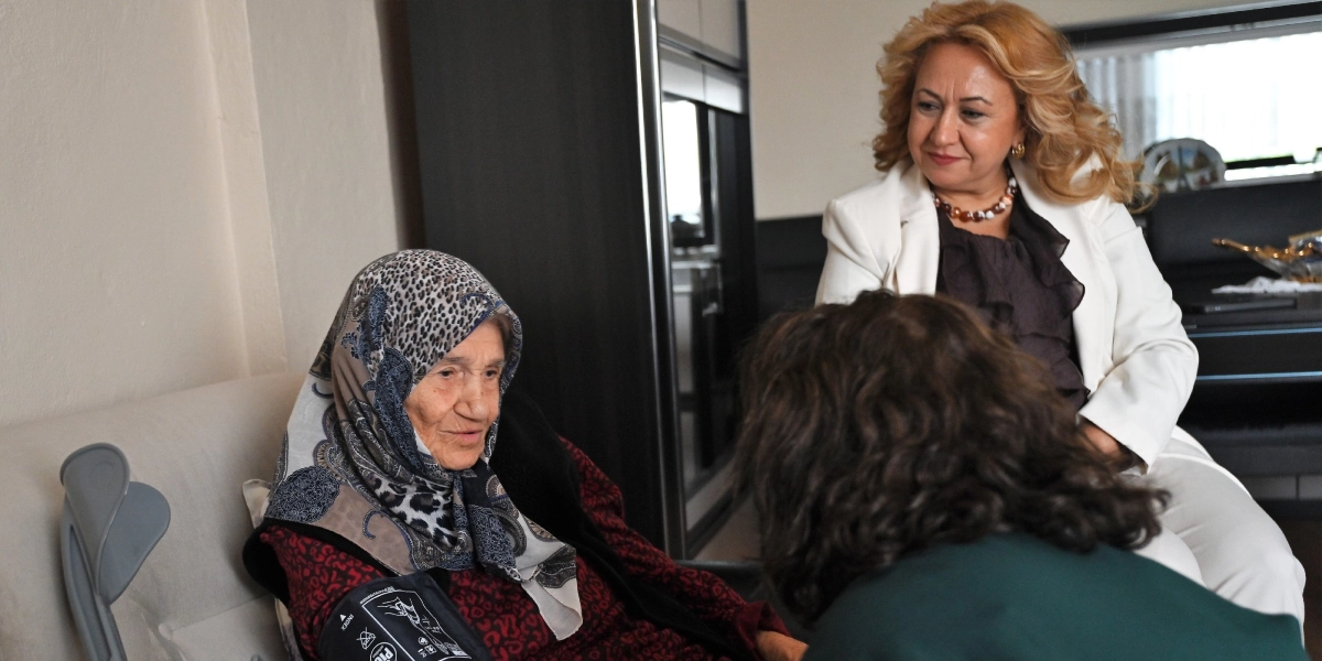 Beyaz ceketli sarışın bir kadın, koltukta oturan ve kolunda tansiyon aleti takılı olan yaşlı bir kadını gülümseyerek izliyor. Yaşlı kadının yanında, sırtı dönük şekilde oturan başka bir kadın ise onunla ilgileniyor.