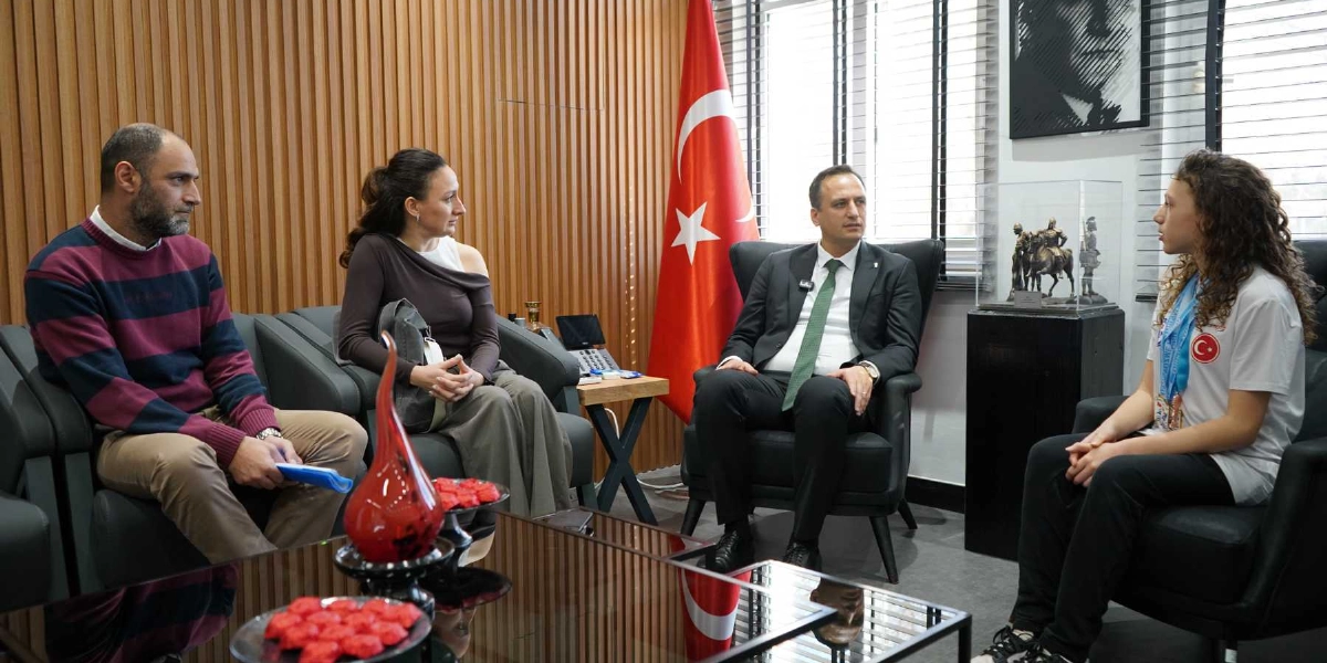 Bornova Belediye Başkanı Mustafa İduğ, makamında boynunda madalyalarla oturan genç bir sporcuyu ve beraberindeki iki yetişkini ağırlıyor. Oda içerisinde Türk bayrağı, Atatürk portresi ve şövale üzerinde bir heykel figürü bulunuyor. Masanın üzerinde kırmızı süs eşyaları yer alıyor.