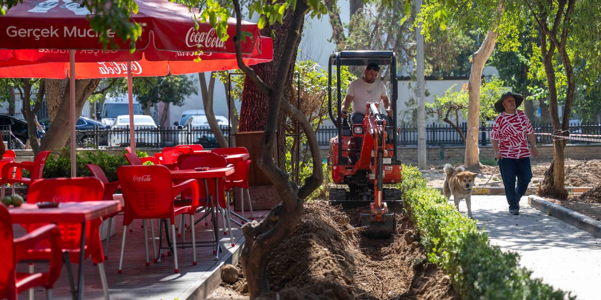 Büyükpark'ta bir operatör, küçük bir iş makinesiyle yeşil alanın kenarında sulama sistemi için olduğu tahmin edilen bir kanal kazıyor. Çalışma alanının hemen yanında kırmızı masa ve sandalyelerden oluşan bir kafe bölümü, diğer tarafta ise köpeğini gezdiren şapkalı bir vatandaş bulunuyor.