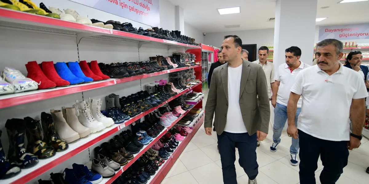 Bornova'da sosyal yardım amaçlı bir mağazada, raflarda dizili çeşitli ayakkabıları inceleyen bir grup erkek ziyaretçi yürüyor. Önde gri ceketli ve beyaz tişörtlü bir kişi, arkasında ise beyaz tişörtlü ve lacivert pantolonlu diğer ziyaretçiler yer alıyor. Duvarlarda 