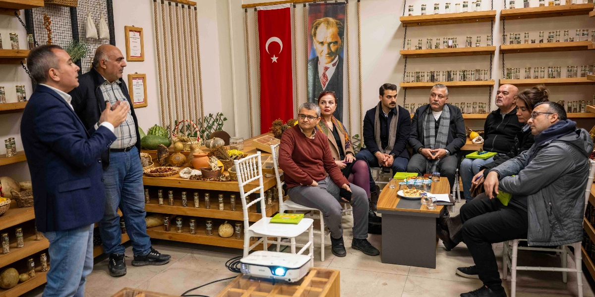 Kapalı bir mekanda, onarıcı tarım üzerine yapılan bir toplantıda, ayakta duran bir konuşmacı karşısında oturan bir grup katılımcı yer alıyor. Arka planda duvarda asılı Türk bayrağı ve Mustafa Kemal Atatürk portresi bulunuyor. Mekanın duvarlarında raflara dizilmiş çok sayıda küçük cam kavanoz, masanın üzerinde ise çeşitli tarım ürünleri ve bir projeksiyon cihazı görülüyor.