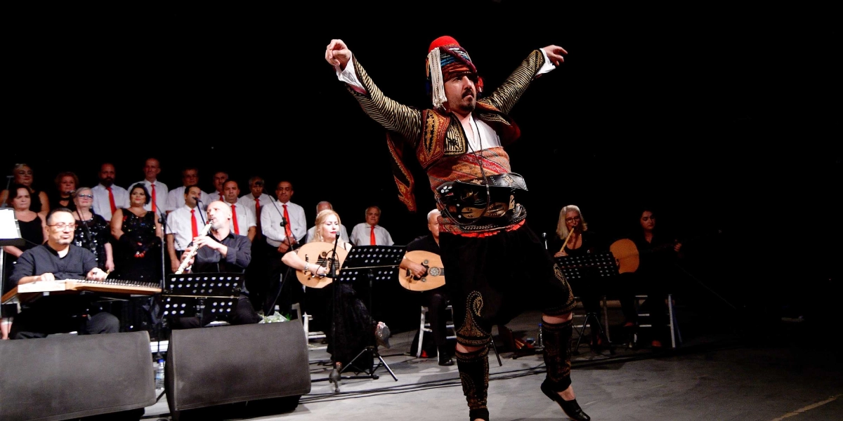 Sahne ışıkları altında, geleneksel Osmanlı kıyafetleri ve başında fesiyle dans eden bir erkek figürü, kollarını iki yana açmış şekilde performans sergiliyor. Arka planda ise beyaz gömlek ve kırmızı kravat giymiş erkekler ile siyah kıyafetli kadınlardan oluşan bir koro ve ud, kanun gibi enstrümanları çalan müzisyenler yer alıyor.