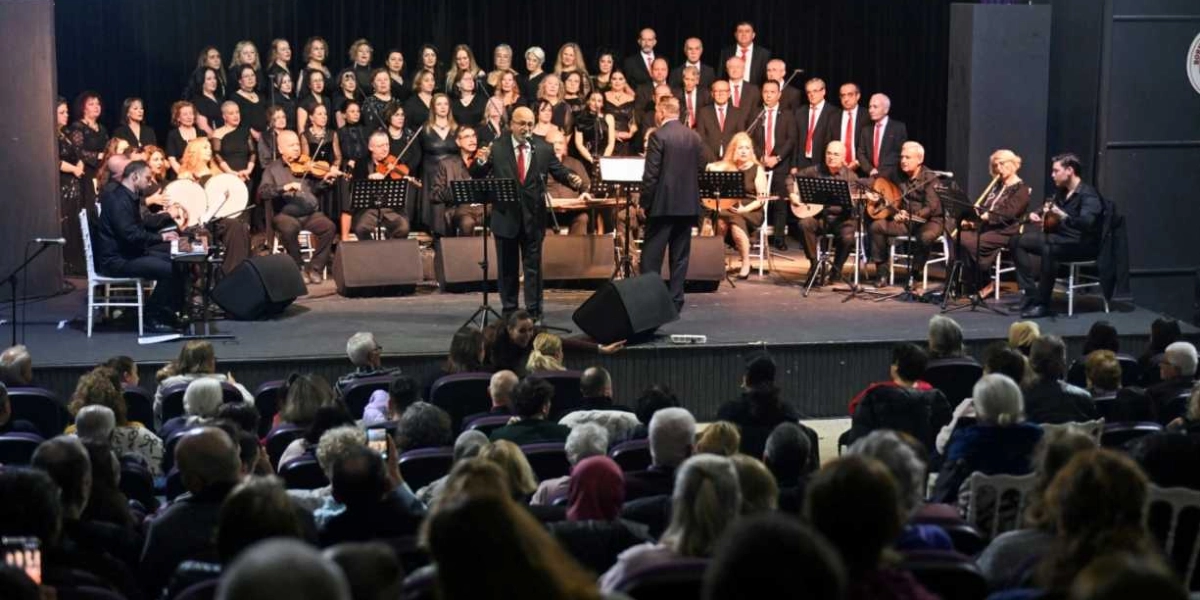 Sahne üzerinde siyah kıyafetler giymiş kalabalık bir koro, orkestra ve solistlerin yer aldığı bir konser performansı. Sahnenin ön kısmında bir orkestra şefi ve mikrofon başında şarkı söyleyen bir solist bulunurken, arkalarında geniş bir koro grubu ve çeşitli müzik aletleri çalan müzisyenler yer alıyor. Sahne, loş ışıklandırılmış bir konser salonunda, dolu bir izleyici kitlesinin önünde gerçekleşiyor.