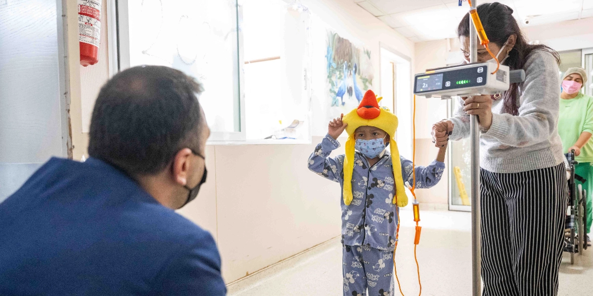 Hastane koridorunda, üzerinde Mickey Mouse desenli pijamaları ve başında sarı bir kuş figürlü şapkası olan maskeli bir çocuk, yanında serum askısını tutan bir yetişkinle birlikte yürüyor. Çocuğun karşısında, ona doğru dönmüş şekilde duran lacivert ceketli bir adam bulunuyor. Arka planda ise tekerlekli sandalyede oturan başka bir kişi daha görülüyor.