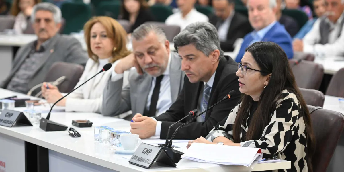 Bornova Belediye Meclisi toplantısında, ön planda mikrofon başında konuşan gözlüklü bir kadın üye ve yanında oturan diğer meclis üyeleri yer alıyor. Masanın üzerinde 