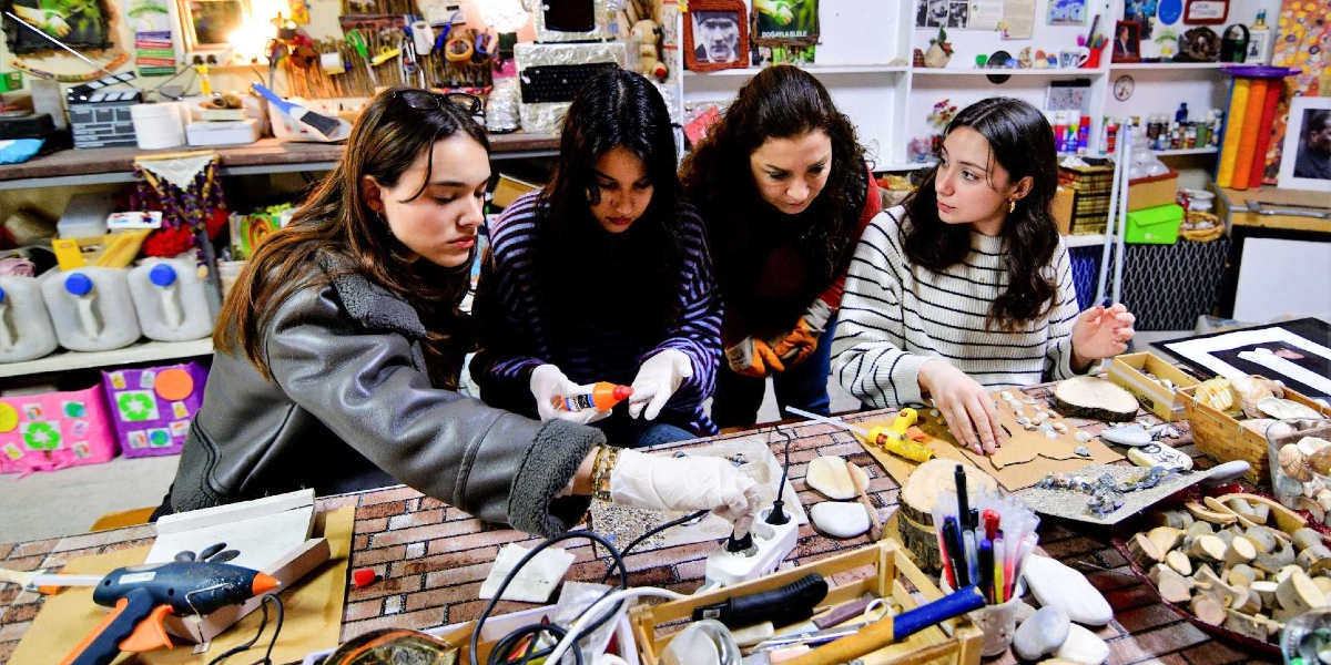 Dört genç kadın, atölye ortamında masanın etrafında toplanmış, çeşitli atık malzemeleri kullanarak sanat eserleri tasarlıyor. Masanın üzerinde silikon tabancaları, ahşap parçaları, deniz kabukları ve boya kalemleri bulunuyor. Gençlerden biri elindeki yapıştırıcıyı kullanırken, diğerleri dikkatle çalışmayı izliyor veya malzemeleri düzenliyor. Arka planda raflarda çeşitli el işi malzemeleri, geri dönüşüm temalı görseller ve Atatürk portresi yer alıyor.