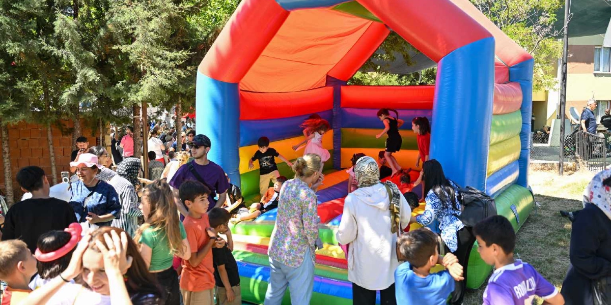 Açık havada kurulan renkli, şişme bir oyun parkında çocuklar zıplayarak eğlenirken, etraflarında onları izleyen aileler ve kalabalık bir grup bulunuyor. Yeşil ağaçların gölgesindeki alanda, şişme oyun alanının önünde duran yetişkinler ve çocuklar şenlikli bir atmosferde vakit geçiriyor.