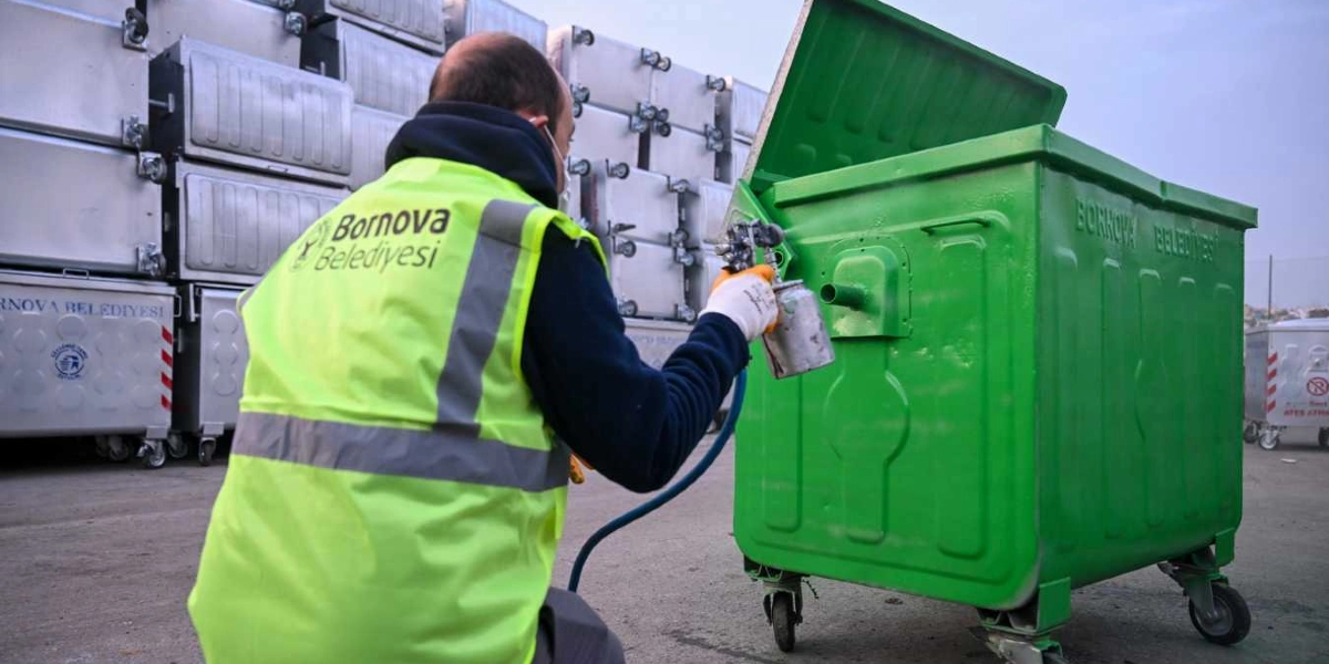 Bornova Belediyesi logolu fosforlu sarı yelek giyen bir belediye çalışanı, elindeki boya tabancasıyla metal bir çöp konteynerini yeşile boyuyor. Arka planda üst üste dizilmiş çok sayıda gri renkli boş çöp konteyneri bulunuyor.