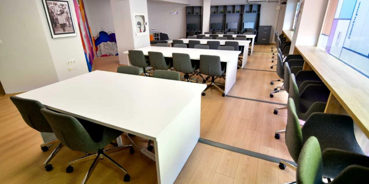 Modern bir çalışma alanı veya kütüphane düzeninde, beyaz masalar ve gri tekerlekli sandalyelerden oluşan oturma düzeni. Odanın bir tarafında pencere kenarı boyunca uzanan ahşap bir çalışma tezgahı, diğer tarafında ise grup çalışmasına uygun dikdörtgen masalar bulunuyor. Arka planda bilgisayar istasyonları ve duvarda Atatürk'ün yer aldığı çerçeveli bir fotoğraf dikkat çekiyor. Zemin açık renkli ahşap parke ile kaplı ve ortam aydınlık bir görünüme sahip.
