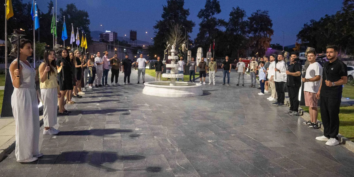 Alacakaranlıkta, Bornova'daki Abide-i Hürriyet anıtının önünde iki sıra halinde dizilmiş bir grup insan, ellerinde yanan meşalelerle duruyor. Merkezde beyaz mermerden bir süs havuzunun bulunduğu taş döşeli meydanda, kadınlar ve erkeklerden oluşan katılımcılar törensel bir duruş sergiliyor. Arka planda ise direklerde asılı çeşitli bayraklar ve ağaçlar görülüyor.