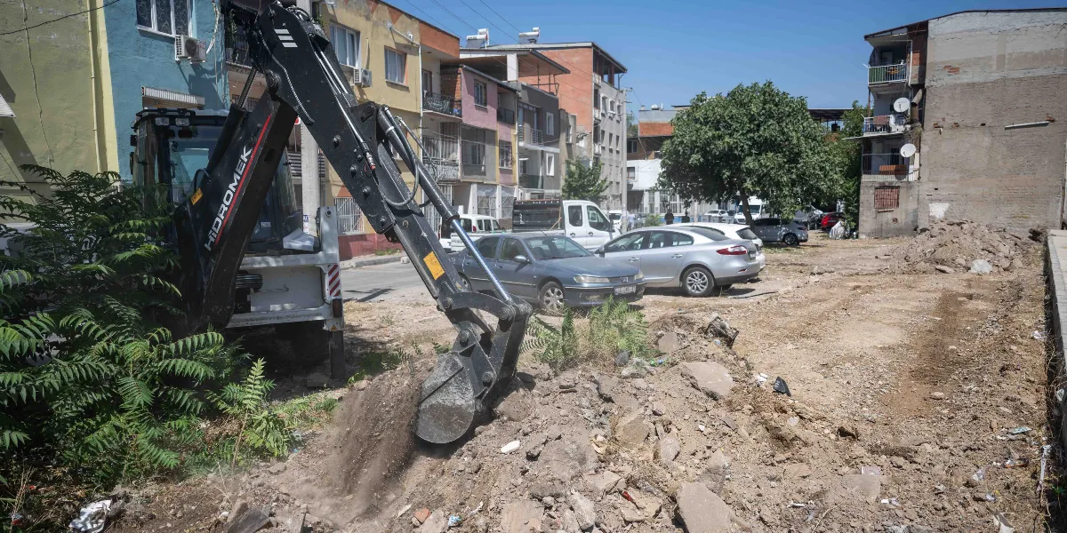 Bornova'da bir sokak arasında, Hidromek marka bir iş makinesi toprak zeminde kazı çalışması yapıyor. Arka planda çok katlı apartmanlar, park halindeki otomobiller ve yeşil bir ağaç görülüyor.