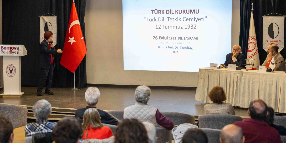 Bir konferans salonunda düzenlenen etkinlikte, kürsüde konuşma yapan bir kadın ve masada oturan iki konuşmacı yer alıyor. Sahnenin arka planındaki yansıtıcı perdede 