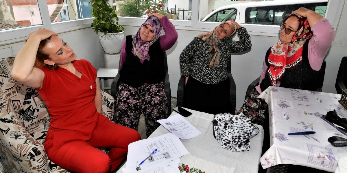 Kırmızı üniformalı bir sağlık çalışanı, başörtülü üç kadına oturarak boyun esnetme egzersizi yaptırıyor. Kadınlar da sağlık çalışanıyla birlikte ellerini başlarının üzerine koyarak aynı esnetme hareketini uyguluyor. Önlerindeki masanın üzerinde egzersiz talimatlarının yer aldığı kağıtlar ve bir kalem bulunuyor.