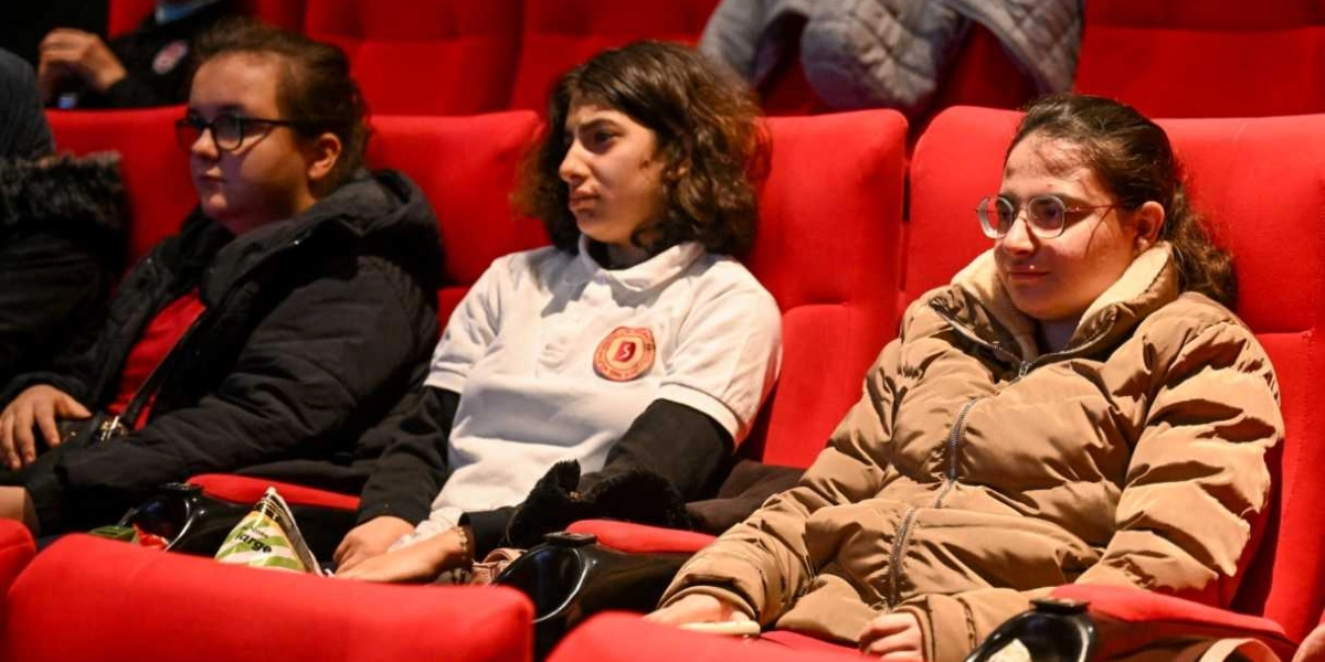 Sinema salonundaki kırmızı koltuklarda yan yana oturan üç genç kız, dikkatle karşıya bakarak film izliyor. En sağdaki kız bej rengi şişme bir mont ve gözlük takarken, ortadaki kız üzerinde okul arması bulunan beyaz bir polo yaka tişört giyiyor. En soldaki kız ise siyah bir mont ve gözlükle oturuyor.