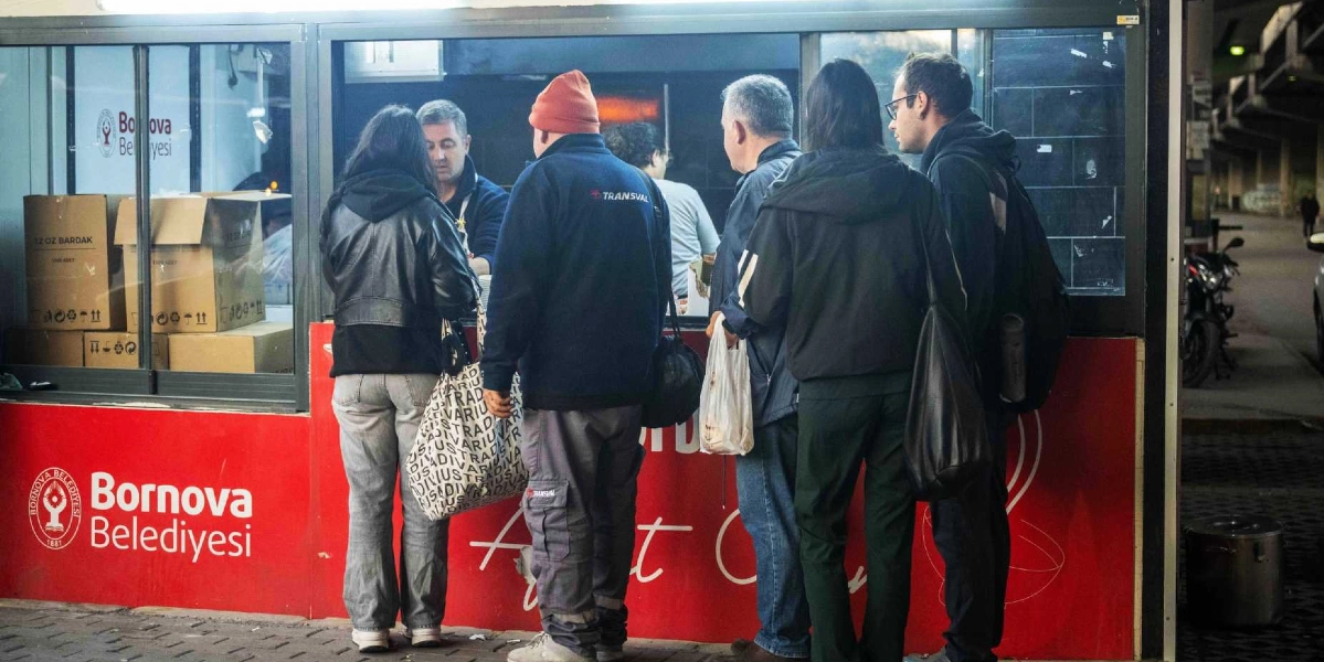 Bornova Belediyesi'ne ait kırmızı renkli bir çorba dağıtım noktasının önünde, sıcak çorba almak için bekleyen bir grup insan. İçeride görevli bir kişi hizmet verirken, dışarıda kışlık kıyafetli vatandaşlar sırayla bekliyor. Dağıtım noktasının camlı bölmesinin içinde karton bardak kolileri görülüyor.