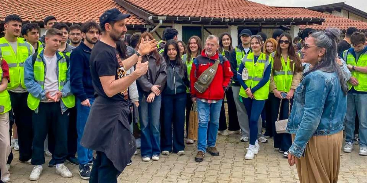 Kiremit çatılı bir yapının önünde toplanmış, çoğunluğu gençlerden oluşan bir grup insan, siyah tişörtlü ve şapkalı bir adamı dinliyor. Gruptaki bazı kişiler, çevre bilincini veya gönüllü çalışmayı simgeleyen parlak sarı reflektörlü yelekler giyiyor. Ön planda, sırtı dönük duran adam el hareketleriyle bir şeyler anlatırken, karşısında gri saçlı, kot ceketli bir kadın ve dikkatle dinleyen kalabalık bir grup yer alıyor. Ortam, açık havada yapılan bir bilgilendirme veya etkinlik başlangıcını yansıtıyor.