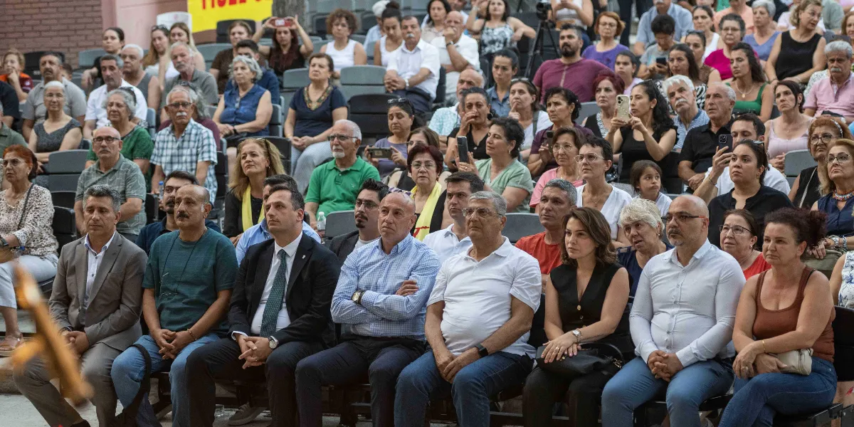 Açık havada, amfi düzenindeki oturma alanında bir araya gelmiş, dikkatle dinleyen kalabalık bir grup insan. Ön sırada takım elbiseli ve günlük kıyafetli kişiler yan yana otururken, arka sıralarda farklı yaş gruplarından katılımcılar yer alıyor. Bazı katılımcılar ellerindeki telefonlarla görüntü alırken, genel atmosfer ciddi ve hüzünlü bir anma törenini yansıtıyor.