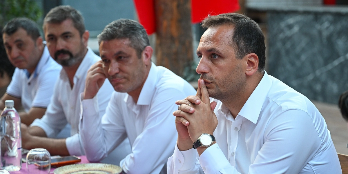 Beyaz gömlek giymiş dört erkek yan yana oturmuş, ciddiyetle bir toplantıyı takip ediyor. En önde oturan kişi ellerini çenesinin altında birleştirmiş, dikkatle karşıya bakıyor. Masanın üzerinde su şişesi ve tabak bulunuyor.