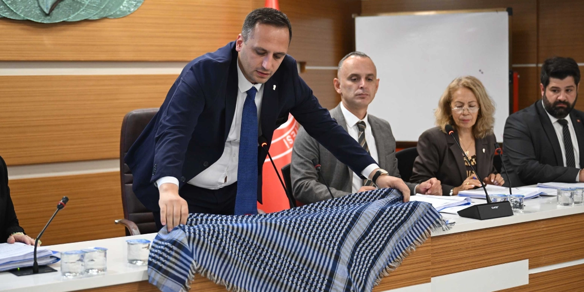 Bornova Belediye Meclis toplantısında, lacivert takım elbiseli ve kravatlı bir yetkili, üzerinde Filistin'i simgeleyen mavi beyaz çizgili bir şalın bulunduğu kürsüde konuşma yapıyor. Masanın devamında oturan diğer meclis üyeleri kendisini dinliyor.
