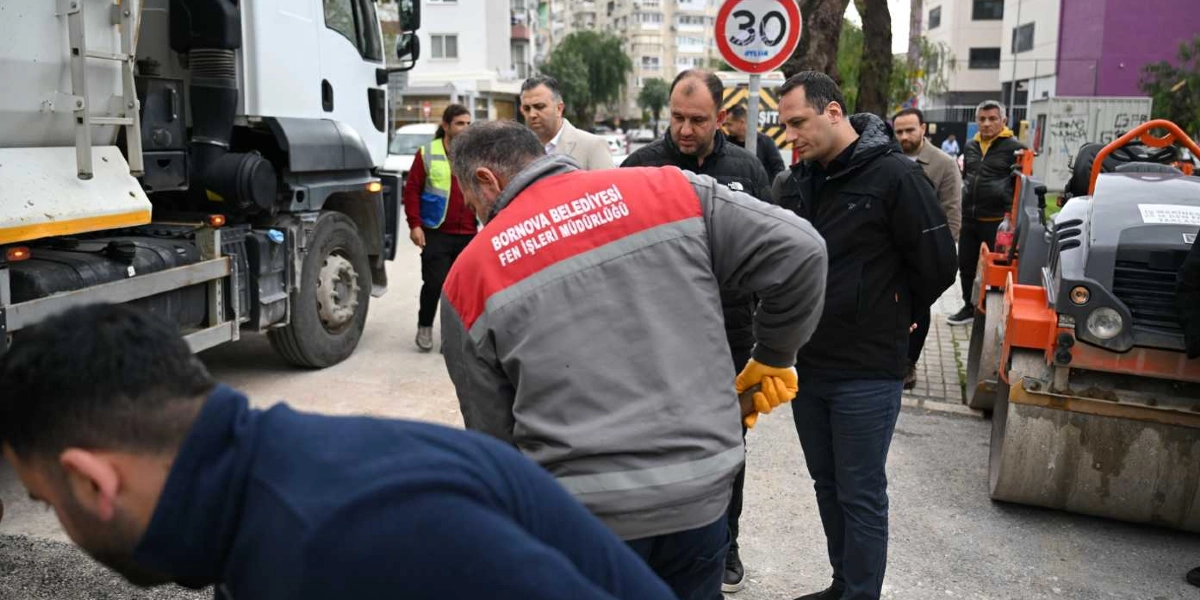 Bornova Belediyesi Fen İşleri Müdürlüğü üniformalı bir çalışan, yol yenileme çalışması sırasında elinde bir aletle eğilmiş vaziyette dururken, çevresindeki yetkililer ve çalışanlar yapılan işi izliyor. Arka planda beyaz bir kamyon, turuncu renkli bir silindir iş makinesi ve 30 kilometre hız sınırını gösteren bir trafik levhası bulunuyor.