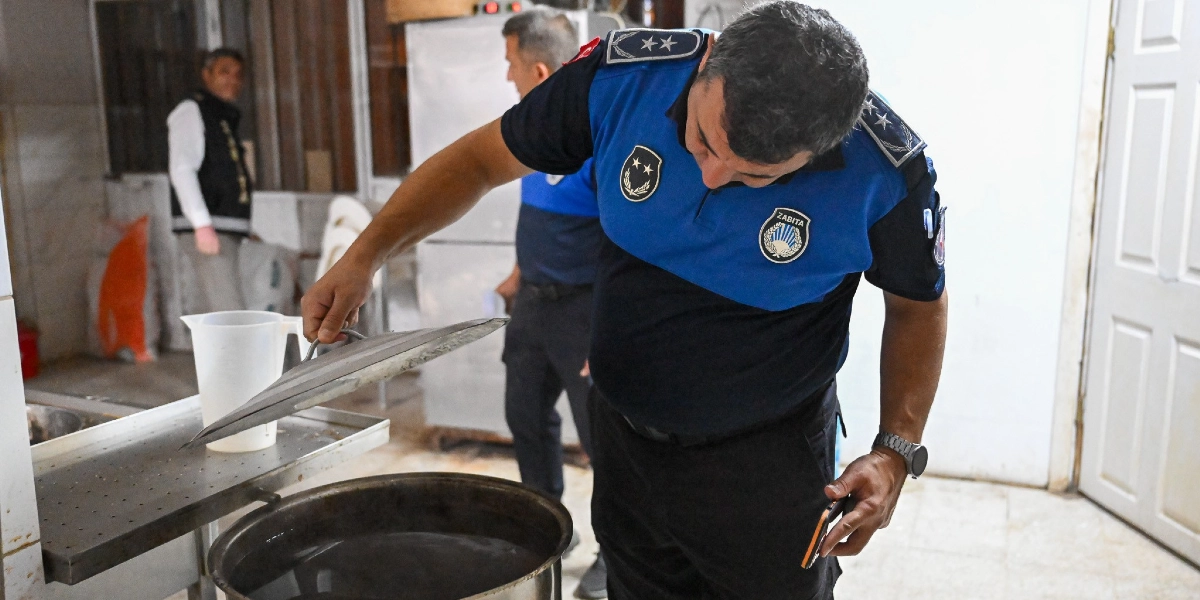 Mavi ve siyah üniformalı bir zabıta memuru, bir fırın veya mutfak ortamında büyük bir tencerenin kapağını kaldırarak içini kontrol ediyor. Arka planda başka bir zabıta memuru ve bir çalışan daha bulunuyor.