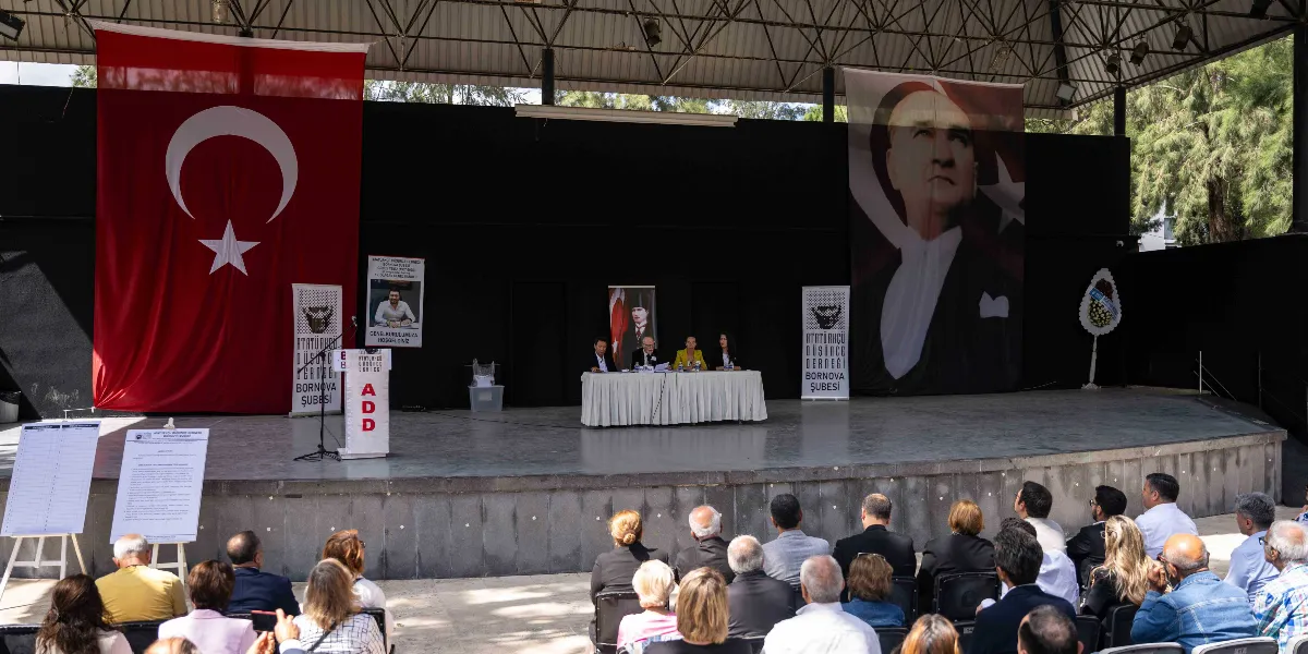 Atatürkçü Düşünce Derneği Bornova Şubesi genel kurul toplantısında, sahnede dört kişilik bir divan heyeti oturuyor. Sahnenin arka planında büyük bir Türk bayrağı, Mustafa Kemal Atatürk'ün portresi ve üzerinde Güney Temiz'in fotoğrafının bulunduğu bir anma pankartı yer alıyor. Sahnenin iki yanında Atatürkçü Düşünce Derneği Bornova Şubesi'ne ait logolu flamalar bulunuyor. Ön planda ise izleyiciler, sahneye dönük şekilde sandalyelerde oturarak toplantıyı takip ediyor.