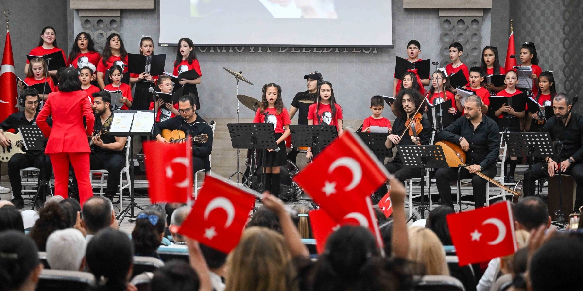 Kırmızı tişörtlü çocuklardan oluşan bir koro ve onlara eşlik eden müzisyenlerin sahne aldığı bir etkinlik. Koro üyeleri ellerinde notalarla şarkı söylerken, önlerinde gitar, keman ve ud çalan müzisyenler yer alıyor. Kırmızı ceketli bir şef koroyu yönetiyor. Sahnenin iki yanında Türk bayrakları bulunuyor. İzleyiciler arasında ellerinde Türk bayrakları sallayan kişiler görülüyor. Arka planda Bornova Belediyesi yazılı bir sahne düzeni yer alıyor.