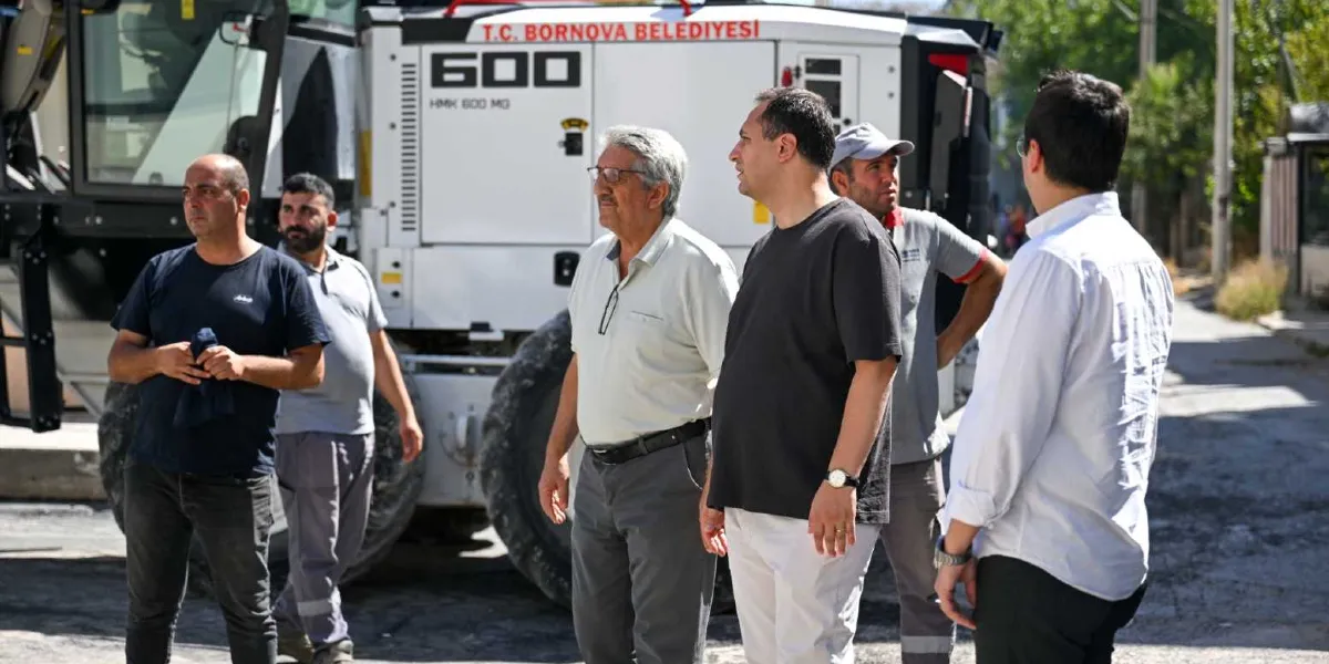 Üzerinde T.C. Bornova Belediyesi yazılı büyük bir iş makinesinin önünde duran, aralarında Bornova Belediye Başkanı Mustafa İduğ'un da bulunduğu bir grup yetkili ve çalışan, devam eden yol çalışmalarını inceliyor.