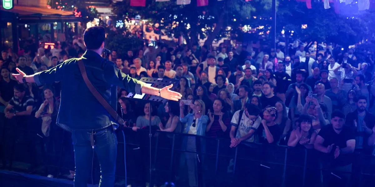 Gece vakti açık havada düzenlenen bir konserde, sahnedeki erkek şarkıcı arkası dönük şekilde kollarını iki yana açarak kalabalığa sesleniyor. Önünde nota sehpası bulunan şarkıcıyı izleyen çok sayıda genç, sahne önündeki bariyerlerin arkasında coşkuyla alkışlıyor ve bazıları telefonlarıyla kayıt alıyor. Sahne ışıklarının mavi tonlarda aydınlattığı konser alanının arka planında ağaçlar ve mekan ışıkları görülüyor.