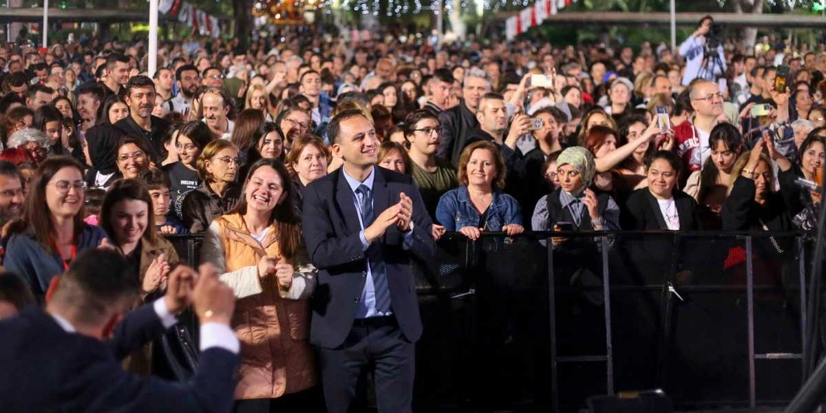 Küçükpark'ta düzenlenen 19 Mayıs gençlik etkinliğinde, sahne önündeki bariyerlerin arkasında toplanmış kalabalık bir grup insan görülüyor. Ön planda, lacivert takım elbiseli ve kravatlı bir erkek alkışlayarak sahneye bakıyor; yanında ise neşeli bir ifadeyle gülen kadınlar ve diğer katılımcılar yer alıyor. Arka planda ise oldukça yoğun bir kalabalık, ellerinde telefonlarla çekim yaparak veya sahneyi izleyerek etkinliğe katılıyor. Ortamda ağaçlar ve ışıklandırmalar dikkat çekiyor.