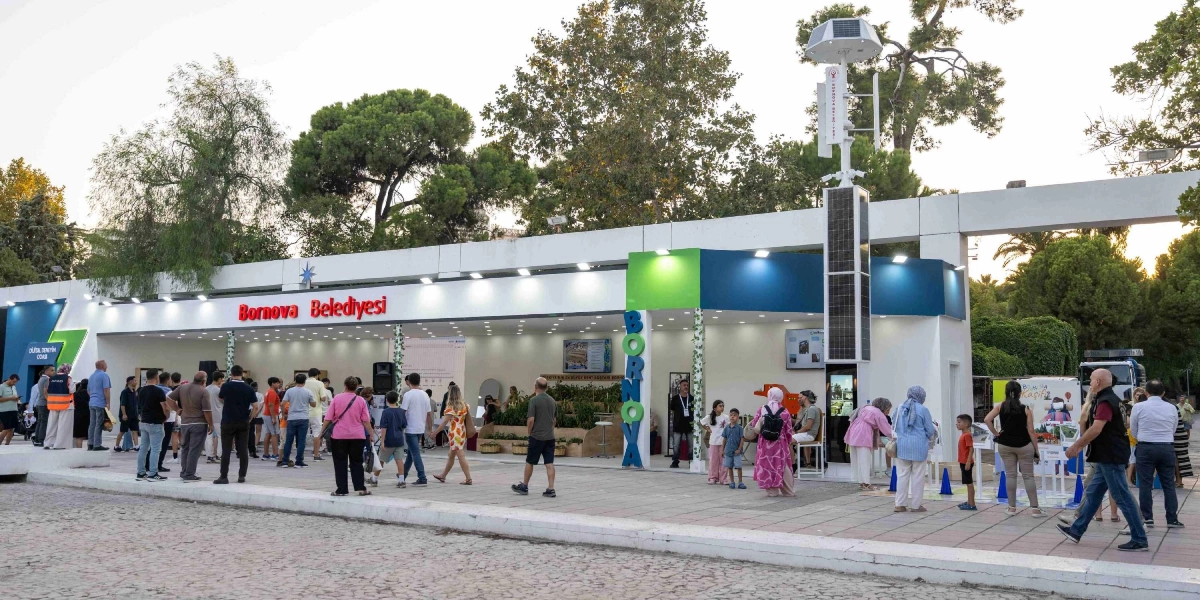 İzmir Enternasyonal Fuarı'ndaki Bornova Belediyesi standının önünde, ziyaretçilerin gezindiği ve incelemelerde bulunduğu açık alan. Beyaz ve modern tasarımlı standın üzerinde kırmızı harflerle Bornova Belediyesi yazısı, yanında ise dikey olarak yerleştirilmiş büyük harflerle Bornova yazısı bulunuyor. Standın önünde güneş enerjisi panelleri ve rüzgar türbini içeren teknolojik bir direk yer alıyor. Çevredeki insanlar standı ziyaret ederken, bazıları stant içindeki yeşil alanları ve dijital ekranları inceliyor.