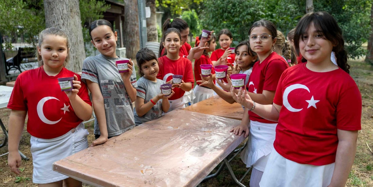 Açık havada, üzerinde Türk bayrağı baskılı kırmızı tişörtler ve beyaz etekler giyen bir grup kız çocuğu, ellerinde 