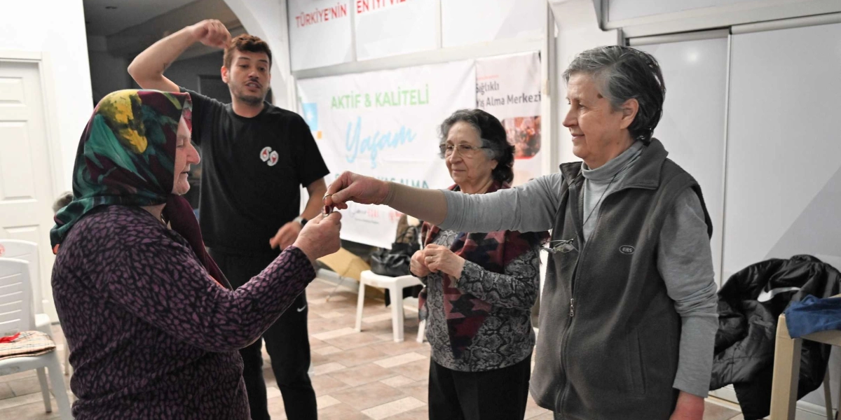 İç mekan bir etkinlik alanında, yaratıcı drama çalışması yapan üç kişi. Ön planda gri yelekli bir kadın, elindeki bir anahtarlığı başörtülü bir kadına uzatıyor. Arkalarında siyah tişörtlü genç bir erkek eğitmen, elleriyle hareketler yaparak süreci yönlendiriyor. Diğer tarafta ise gri desenli bir hırka giymiş yaşlı bir kadın, gülümseyerek onları izliyor. Arka planda üzerinde 