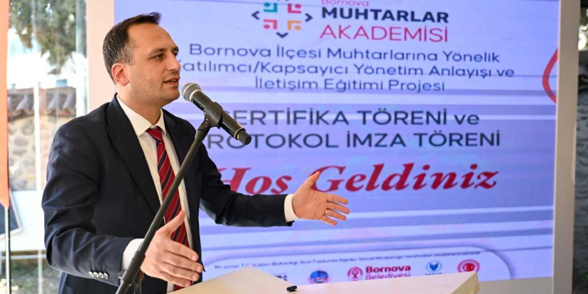 Lacivert takım elbiseli ve çizgili kravatlı bir erkek, kürsüde mikrofon başında konuşma yapıyor. Arkasındaki ekranda 