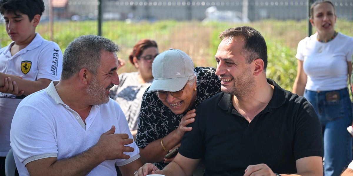 Açık havada, bir tarım alanında bir araya gelmiş neşeli bir grup insan. Ön planda beyaz polo yaka tişörtlü, sakallı bir adam ile siyah polo yaka tişörtlü bir adam birbirlerine bakarak gülümsemektedir. İkisinin arasında, gri şapkalı ve gözlüklü bir kadın da onlara eşlik ederek gülmektedir. Arka planda ise beyaz tişörtlü bir genç ve ayakta duran bir kadın daha yer almaktadır.