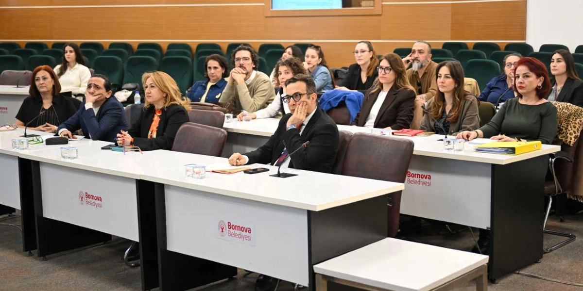 Bornova Belediyesi logolu masaların bulunduğu bir toplantı salonunda, farklı yaş ve meslek gruplarından oluşan katılımcılar sıralarda oturarak dikkatle dinliyor. Katılımcıların bir kısmı masalardaki mikrofonlara yakın dururken, ön planda yer alan kişiler toplantıyı takip ediyor, arka sıralarda ise diğer katılımcılar yer alıyor. Salonun genelinde resmi bir çalışma ortamı hakim.