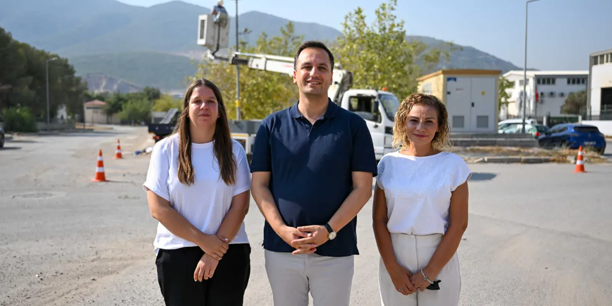 Açık havada, arka planda dağ manzarası ve bir sepetli vinç bulunan asfalt bir alanda yan yana duran üç kişi. Ortada lacivert polo yaka tişörtlü bir erkek, sağında ve solunda ise beyaz bluz giymiş iki kadın gülümseyerek kameraya bakıyor. Çevrede trafik konileri ve bazı teknik yapılar yer alıyor.
