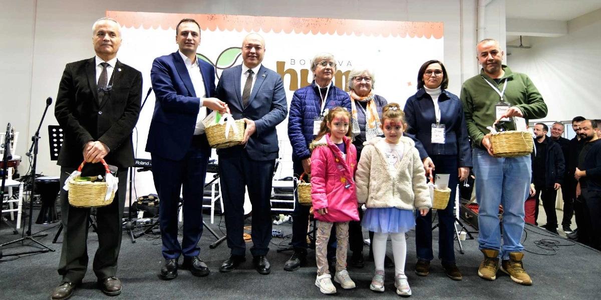 Bornova Tohum Takas Şenliği'nde ellerinde tohum dolu hasır sepetler tutan bir grup yetişkin ve iki çocuk, sahnede yan yana durarak poz veriyor. Arka planda üzerinde 