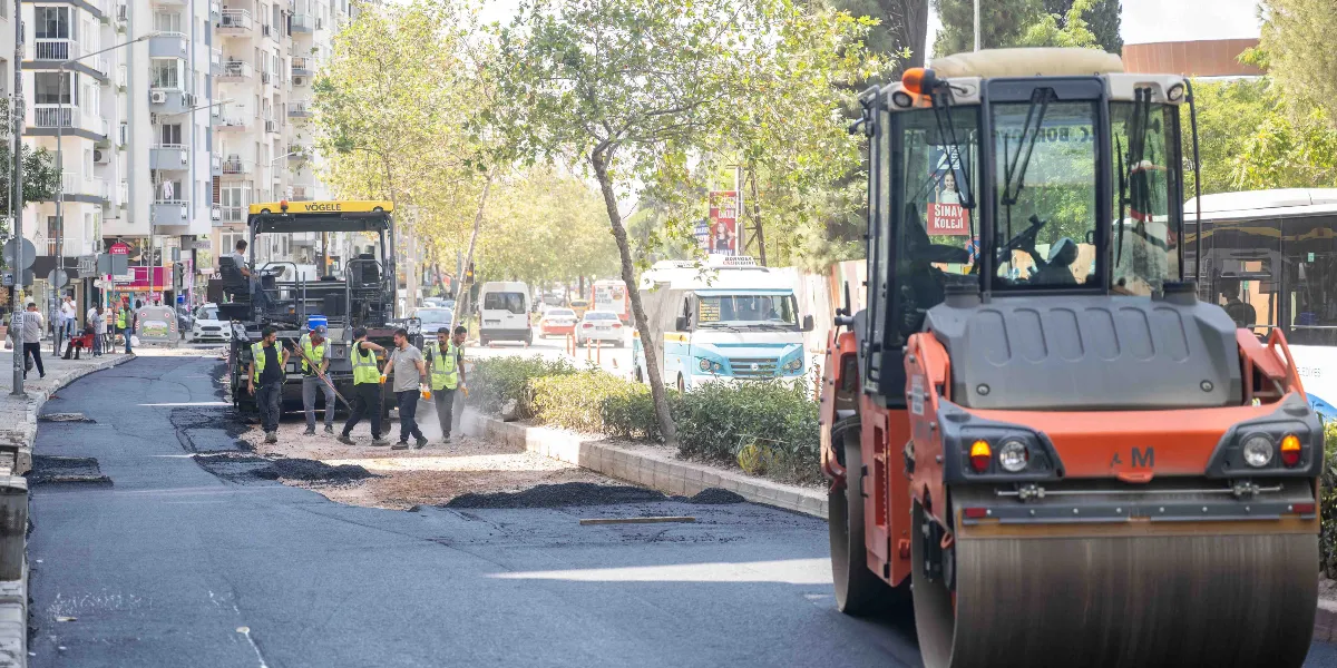 Bornova'da bir cadde üzerinde devam eden yol yenileme çalışmaları kapsamında, reflektörlü iş yeleği giymiş bir grup işçi asfalt serme makinesinin çevresinde çalışıyor. Ön planda turuncu renkli büyük bir silindir iş makinesi yer alırken, arka planda ağaçlıklı bir orta refüj, trafik akışının devam ettiği cadde ve çok katlı binalar görülüyor.