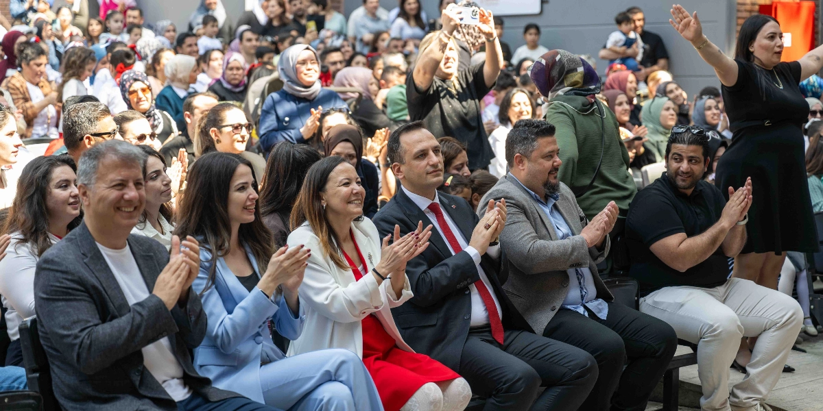 Açık havada düzenlenen bir etkinlikte, ön sırada yan yana oturarak alkışlayan bir grup yetişkin ve arkalarında onları izleyen kalabalık bir izleyici topluluğu. Ön sıradakiler arasında koyu renk ceketli erkekler, açık mavi takım elbiseli bir kadın, beyaz ceket ve kırmızı elbise giyen bir kadın ile siyah tişörtlü bir erkek yer alıyor. Sağ tarafta ise ayakta duran ve kollarını havaya kaldırmış siyah elbiseli bir kadın bulunuyor. İzleyicilerin çoğu gülümseyerek sahnedeki gösteriyi takip ediyor.