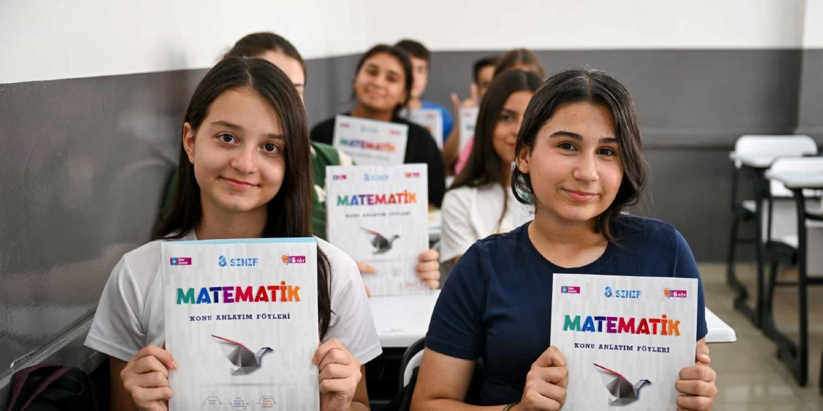 Sınıfta yan yana oturan ve gülümseyerek ellerinde 8. sınıf matematik konu anlatım föylerini tutan iki kız öğrenci ile arka sıralarda oturan diğer öğrenciler.
