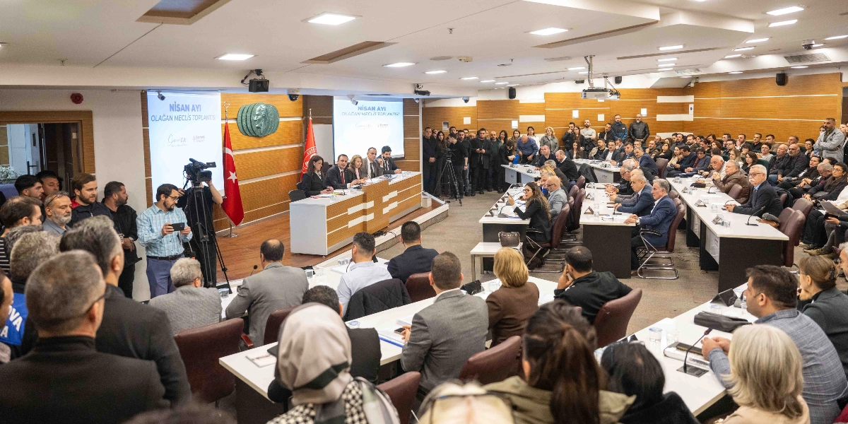 Bornova Belediyesi'nin Nisan ayı olağan meclis toplantısının yapıldığı geniş bir toplantı salonu. Salonun ön kısmındaki kürsüde yetkililer otururken, salonun geri kalanında meclis üyeleri ve izleyiciler sıralar halinde yer alıyor. Arka duvarda üzerinde Atatürk silüeti bulunan kabartma bir pano ve iki yanında 