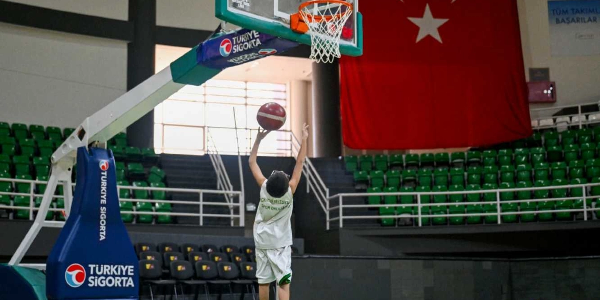 Bornova Belediyesi Spor Okulları forması giyen bir çocuk, kapalı bir spor salonunda basketbol potasına atış yapıyor. Arka planda büyük bir Türk bayrağı ve yeşil koltuklu tribünler yer alıyor. Basketbol potasının üzerinde Türkiye Sigorta logosu bulunuyor.