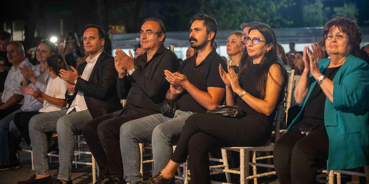Açık havada düzenlenen bir etkinlikte, yan yana dizilmiş sandalyelerde oturan ve alkışlayan bir grup insan. Ön sırada, koyu renkli kıyafetler giymiş erkekler ve kadınlar dikkatle sahneye bakarak alkış tutuyor. Arka planda ise etkinliğe katılan diğer izleyiciler ve hafif loş bir ortam görülüyor.