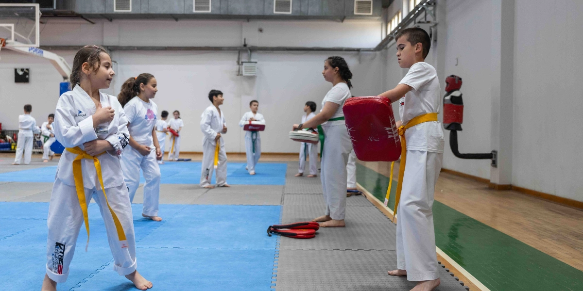 Kapalı bir spor salonunda tekvando antrenmanı yapan çocuklar; ön planda sarı kuşaklı bir kız çocuğu ve karşısında elinde kırmızı bir darbe yastığı tutan sarı kuşaklı bir erkek çocuğu bulunuyor. Arka planda ise beyaz tekvando kıyafetleri giymiş diğer çocuklar farklı eşleşmelerle çalışmaya devam ediyor.