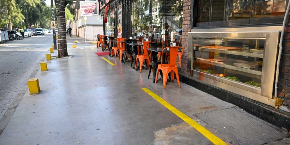 Bornova'da bir cadde üzerinde, işletmelerin kaldırım işgalini önlemek amacıyla yaya yoluna çekilmiş sarı çizgiler ve kaldırım kenarına yerleştirilmiş sarı güvenlik dubaları görülüyor. İşletmenin dış mekanında, sarı çizgilerin arkasında konumlandırılmış turuncu metal sandalyeler ve masalar yer alıyor.